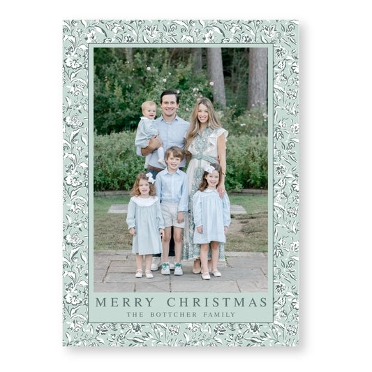 A family of five, consisting of a father, mother, and three children, posing for a photograph in a park setting. The family is dressed in light blue outfits, and the background includes trees and bushes. The image is framed with a floral border, and the text &quot;Merry Christmas&quot; is displayed at the bottom, indicating the holiday season.