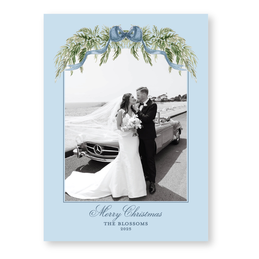 A black and white photo of a bride and groom standing next to a car, with a decorative blue ribbon and greenery frame around the image.