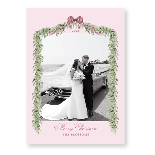 A black and white photo of a bride and groom standing next to a car, framed by a decorative border with greenery and a pink ribbon.