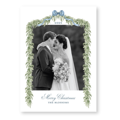 A black and white photograph of a bride and groom embracing, framed by a decorative border with greenery and a blue ribbon. The text "Merry Christmas" and "THE BLOSSOMS" is displayed below the couple.