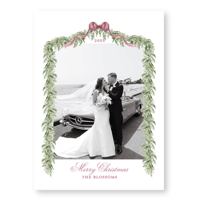 A black and white photograph of a bride and groom standing next to a car, framed by a decorative border with greenery and a pink ribbon. The text at the bottom of the image reads "Merry Christmas THE BLOSSOMS".